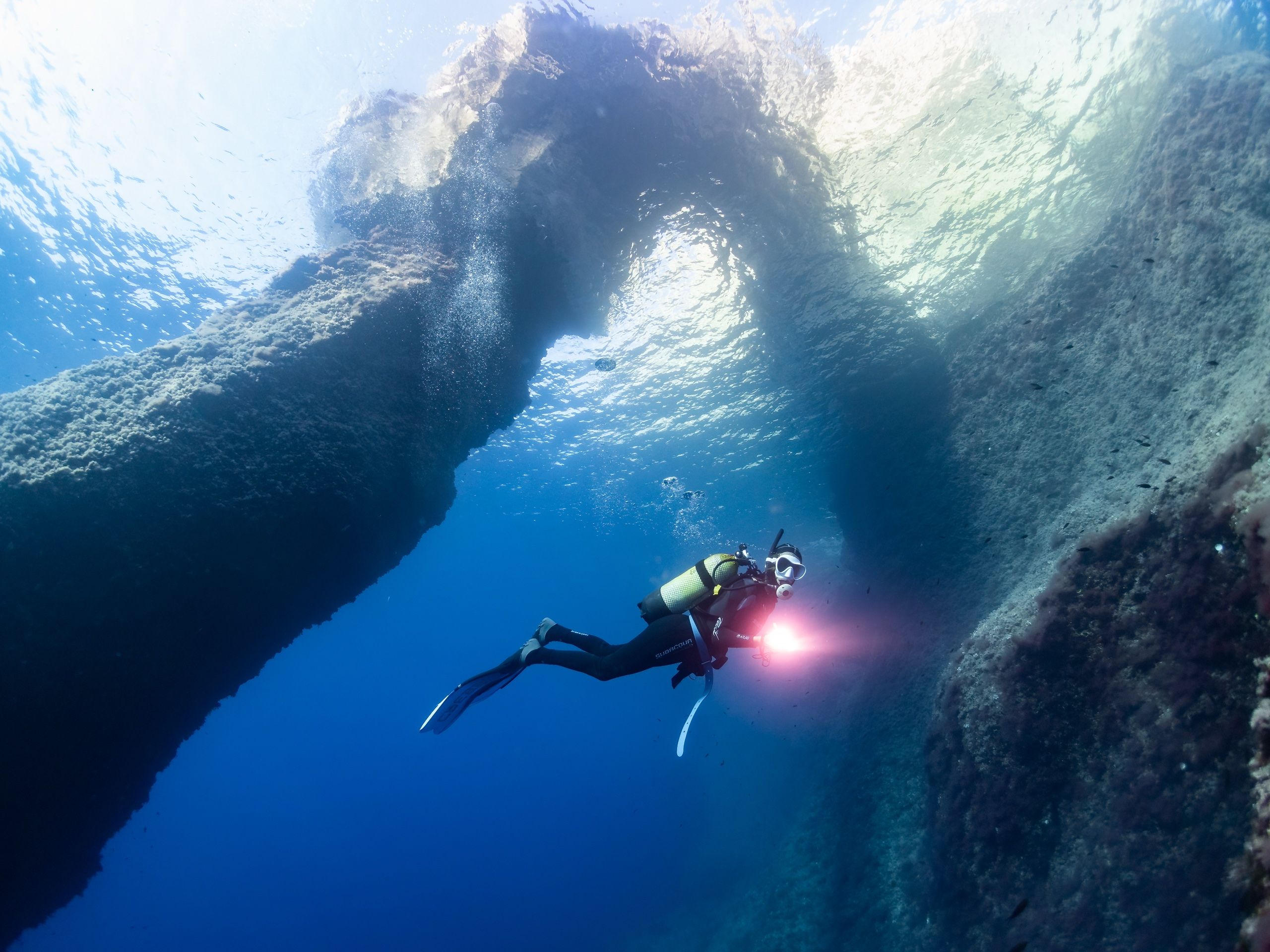 A diver with a lit torch swims under a huge rock arch in deep blue water
